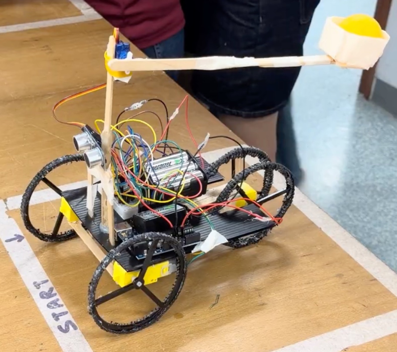The finished robot at the start line: three wheels, a polypropylene base, an ice cream stick arm and a tape cup holding the ball