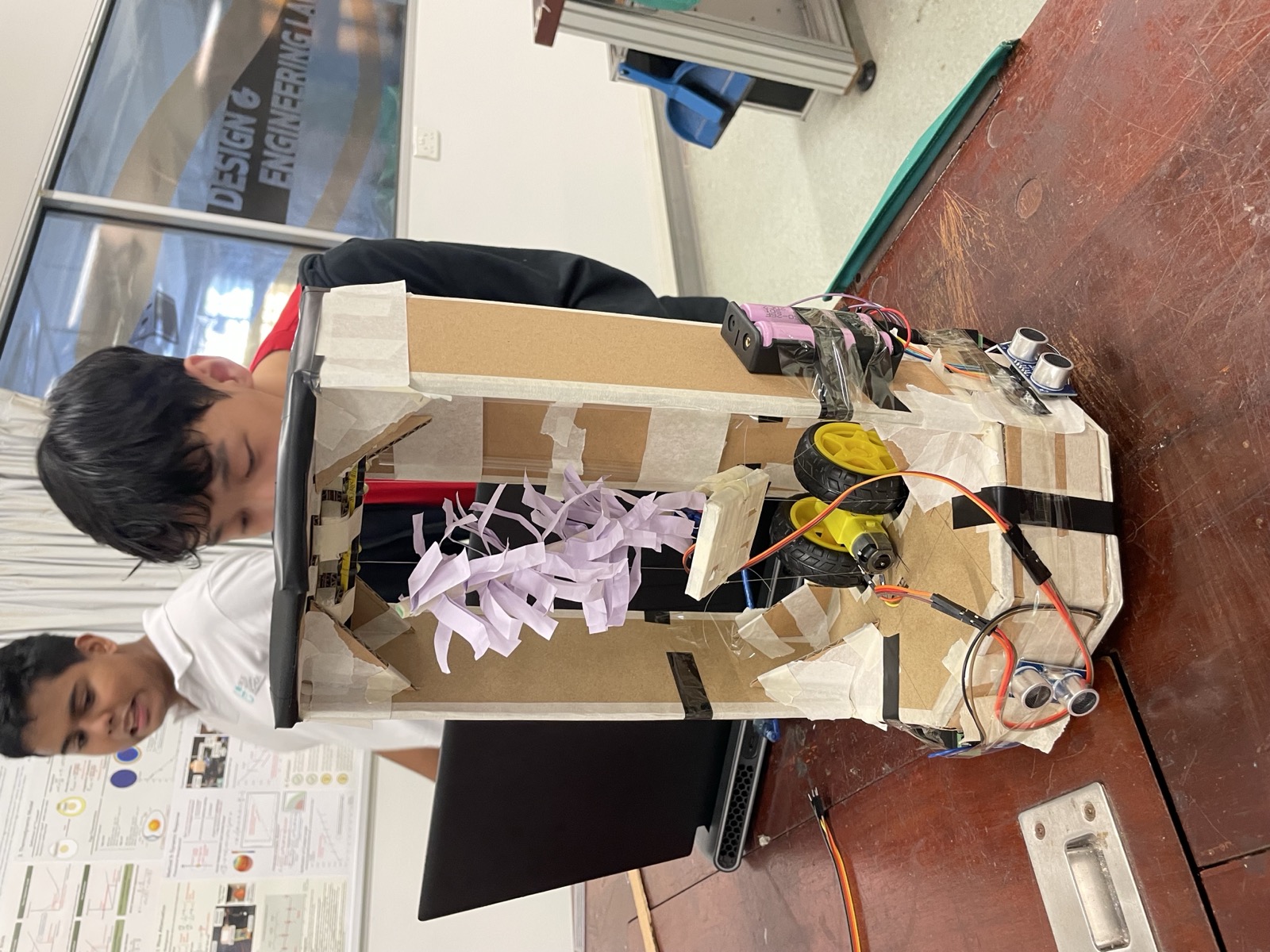 The finished cardboard duster robot with a paper duster hanging inside its frame and ultrasonic sensors at the front
