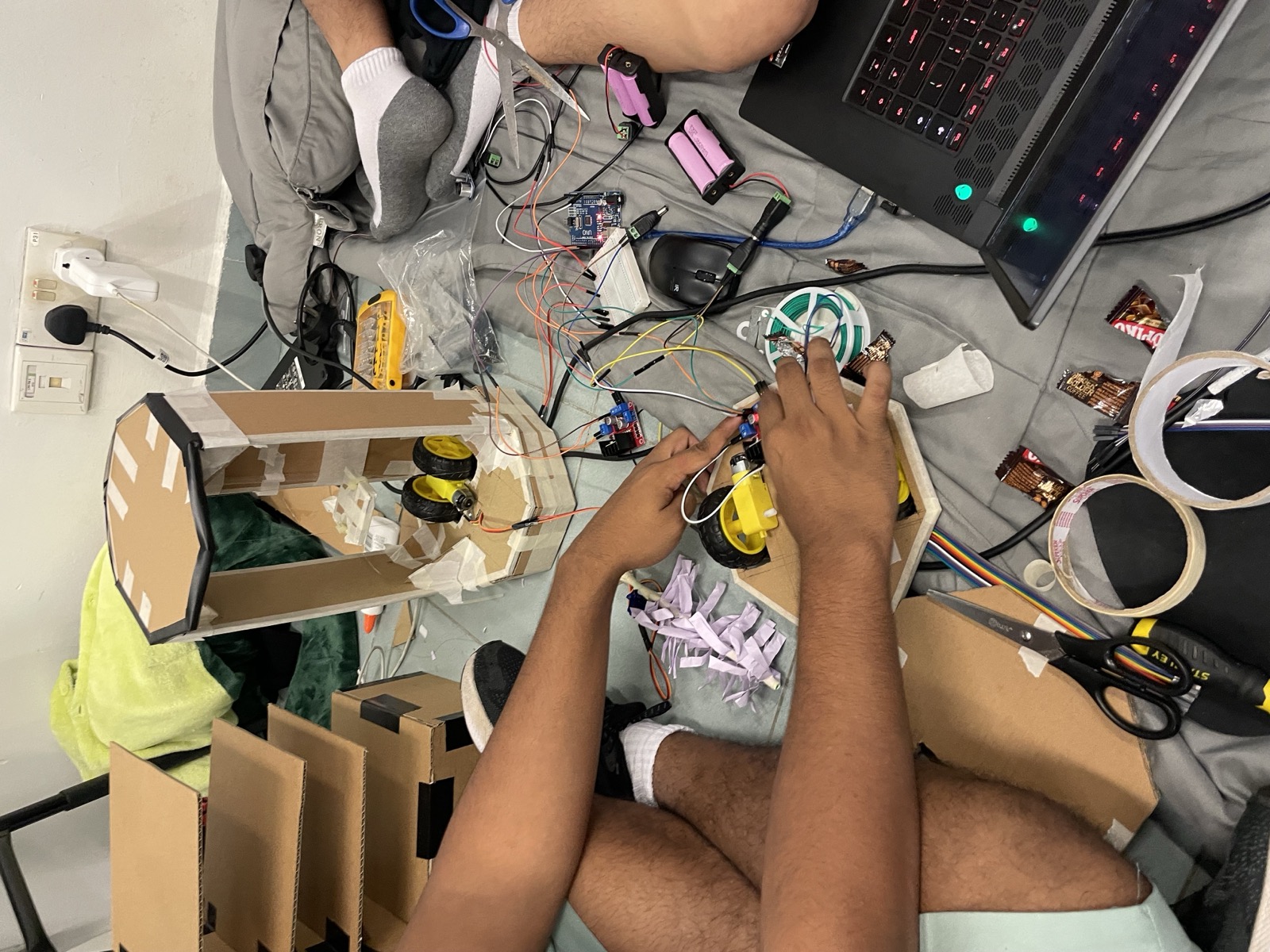 Cardboard frame, wheels, an Arduino and wires spread on the floor mid-build, with snacks and tape around