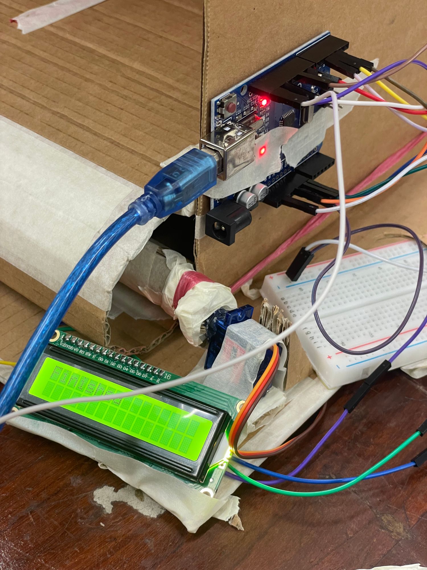 Close-up of an Arduino UNO taped to the side of the cardboard box, wired to a breadboard and a green LCD screen