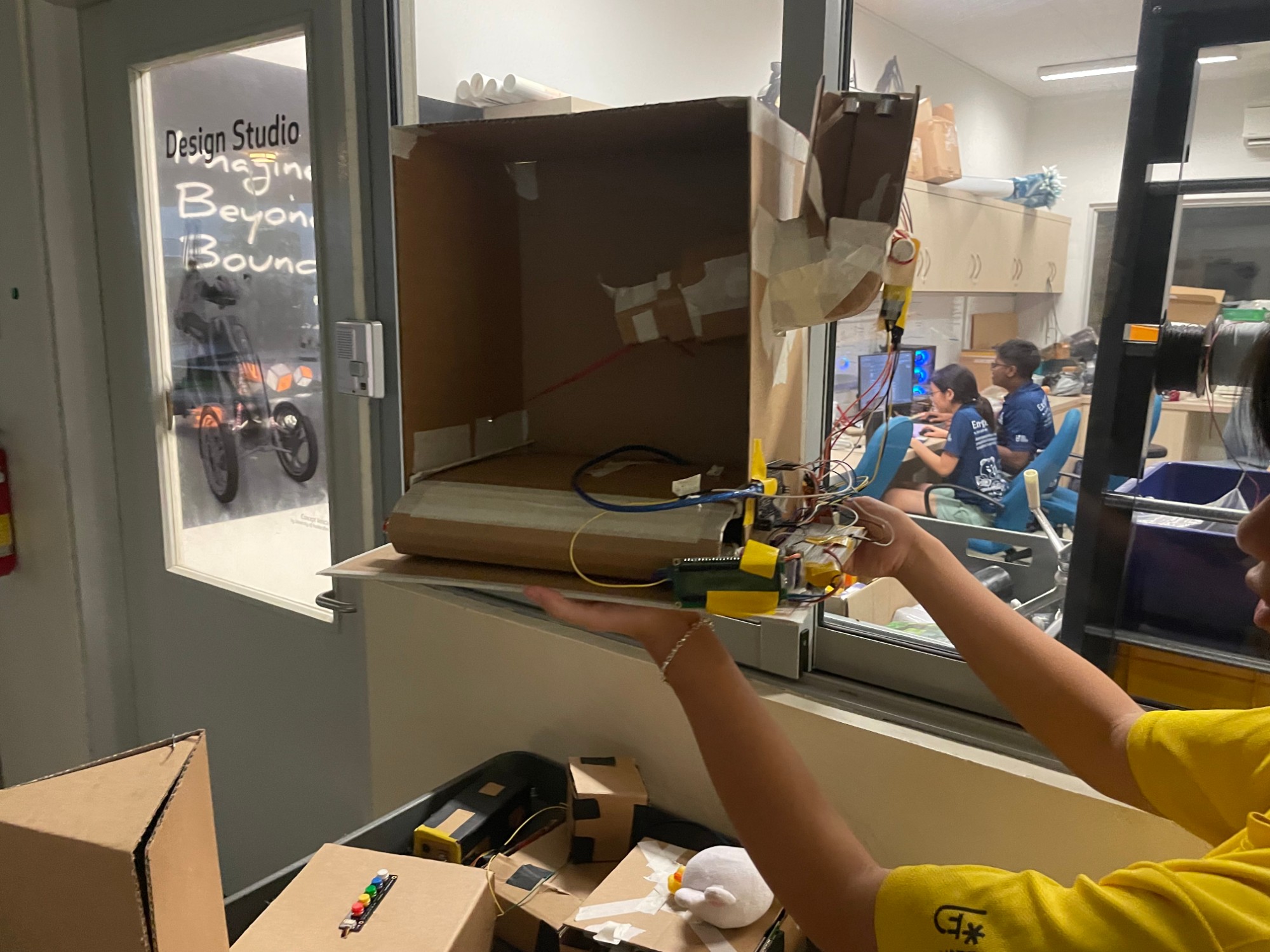 The finished cardboard cage prototype held up in front of the Design Studio window, with the feeder arm on the right and the electronics underneath