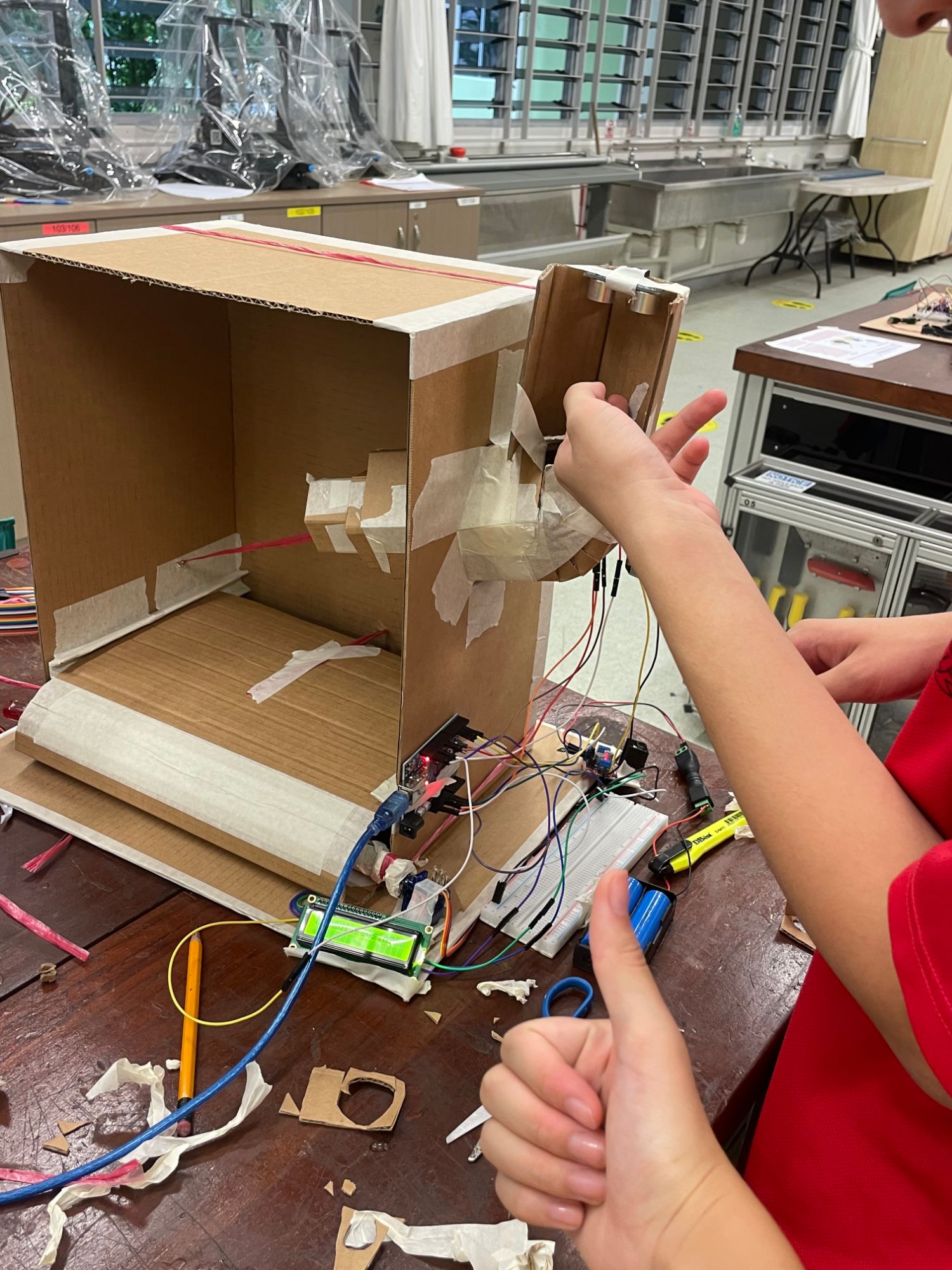 The cardboard cage on a workbench with the rolled-up floor at the front, the feeder arm on the side, and an Arduino, breadboard and LCD wired up below